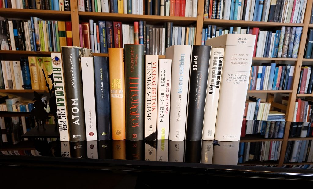 "Stapel von Büchern in einem Regal mit verschiedenen Titeln und Autoren. Von oben nach unten sind folgende Bücher zu sehen:
'Rifleman' von Victor Gregg mit Rick Stroud,
'Karte und Gebiet' von Michel Houellebecq,
'Viking Britain' von Thomas Williams,
'Atom' von Steffen Kopetzky,
´Austerlitz´ von W.G. Sebald,
'Noli me Tangere' von José Rizal,
'Theodoros' von Mircea Cărtărescu,
'Melitta von Stauffenberg' von Thomas Medicus,
'Die Kinder der Schande' von Jean-Paul Picaper und Ludwig Norz,
'Nicht ungeschoren davongekommen' von Ebba D. Drolshagen,
'Geschichte der Völkerwanderung' von Mischa Meier.
Im Hintergrund sind weitere Bücherregale mit einer Vielzahl von Büchern zu sehen.