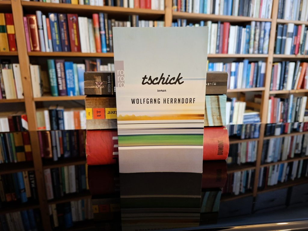 Buchcover von 'Tschick', einem Roman von Wolfgang Herrndorf, das zwischen Bücherregalen in einer Bibliothek oder einem Buchladen steht. Das Cover zeigt eine minimalistische Illustration mit horizontalen Streifen in Grün-, Gelb- und Grautönen. Der Titel 'tschick' ist handschriftartig in Schwarz auf weißem Hintergrund gedruckt