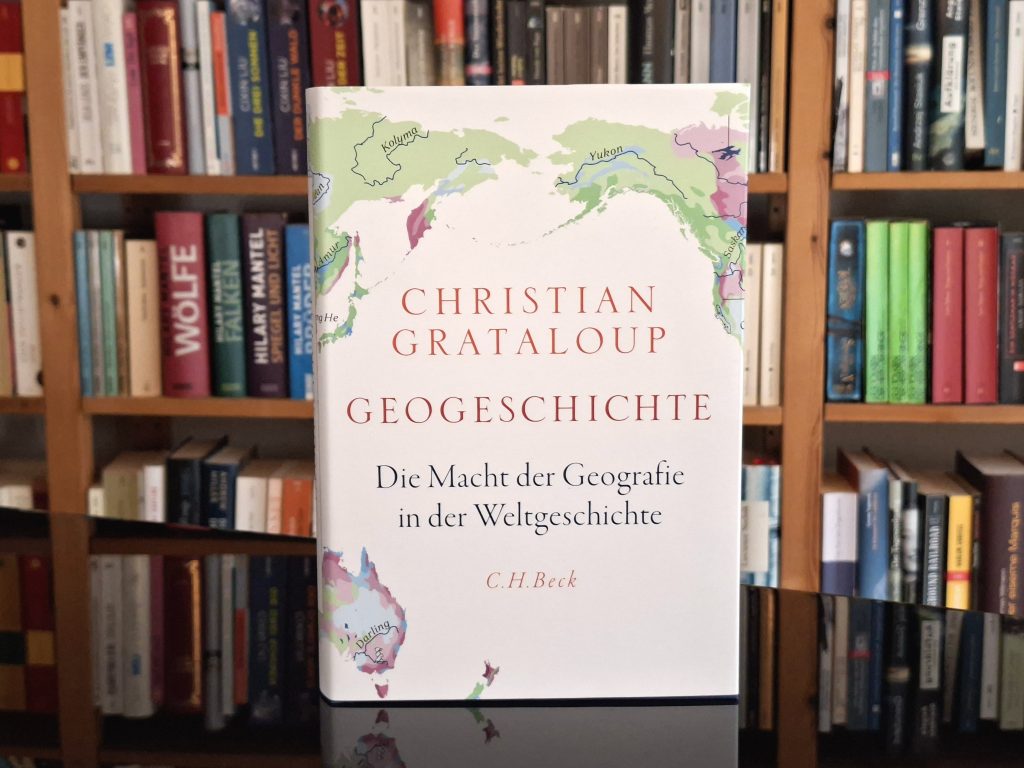 Buchcover von ‚Christian Grataloup: Geogeschichte – Die Macht der Geografie in der Weltgeschichte‘ von C.H.Beck, platziert vor einem Bücherregal mit verschiedenen Büchern.