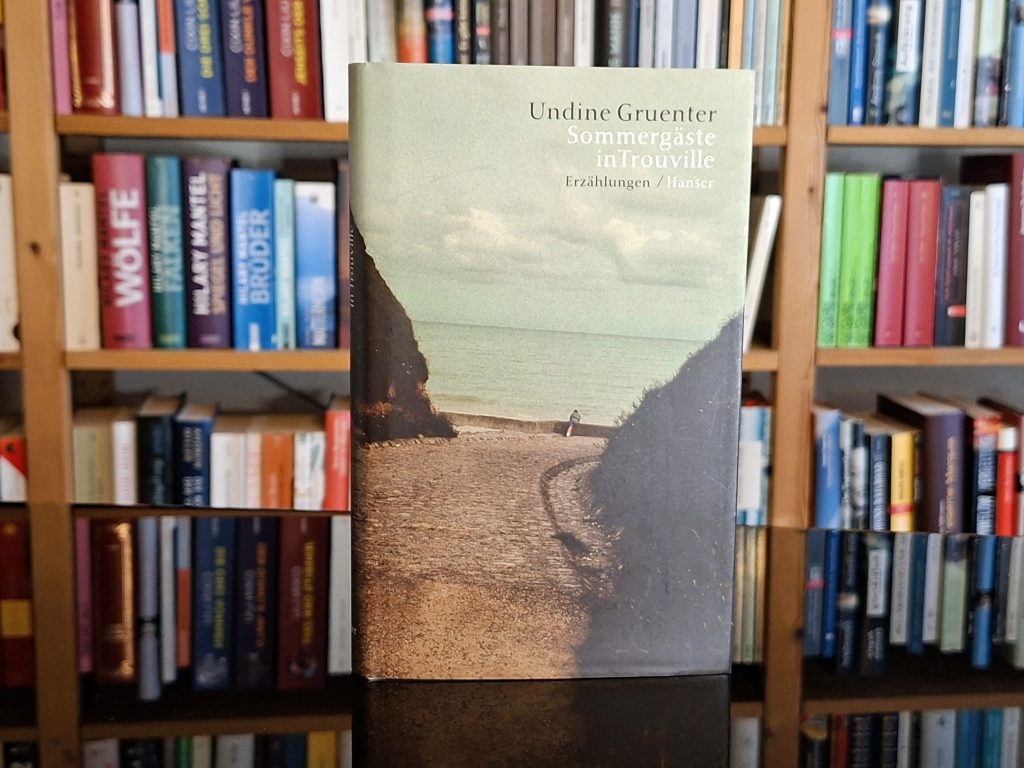 Das Cover des Erzählungsbande "Sommergäste in Trouville" von Undine Gruenter. Eine gepflasterte Straße, das Meer im Hintergrund, ein Junge lehnt an einer Mauer und blickt aufs Wasser.
