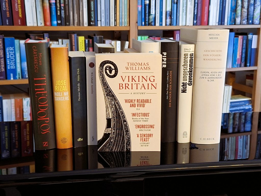 Bücherregal mit einer Auswahl an historischen und literarischen Büchern. Im Vordergrund steht das Buch ‚Viking Britain‘ von Thomas Williams, umgeben von weiteren Werken zu Geschichte, Kultur und Literatur.