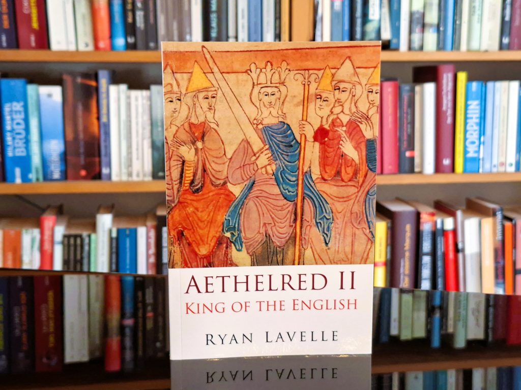 Buchcover von 'Aethelred II: King of the English' von Ryan Lavelle, das vor einem Bücherregal steht. Das Cover zeigt eine mittelalterliche Illustration von gekrönten Personen in traditioneller Kleidung.