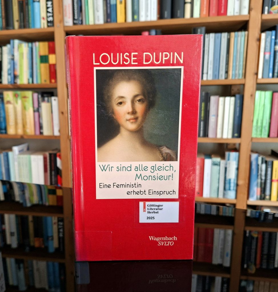 Ein Buch mit dem Titel „Wir sind alle gleich, Monsieur!“ von Louise Dupin steht vor einem gut gefüllten Bücherregal. Der Untertitel lautet „Eine Feministin erhebt Einspruch“. Das Buchcover ist rot mit einem Porträt einer Frau in historischer Kleidung und einem weißen Etikett mit dem Text „Göttinger Literatur Herbst 2025“
