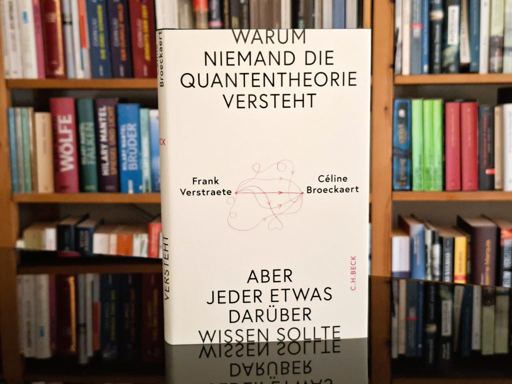 Ein Buch mit dem Titel „Warum niemand die Quantentheorie versteht“ von Frank Verstraete und Céline Broekaert steht vor einem Bücherregal. Der Untertitel lautet „Aber jeder etwas darüber wissen sollte“. Das Buchcover ist hellgrau mit schwarzer Schrift und einer roten Zeichnung von verschlungenen Linien.