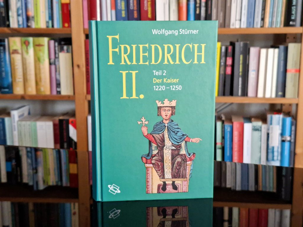 Ein Buch mit dem Titel 'Friedrich II., Teil 2: Der Kaiser 1220–1250' von Wolfgang Stürner, das vor einem Bücherregal steht. Das Cover zeigt eine Illustration von Kaiser Friedrich II. auf einem Thron mit Krone und Zepter.
