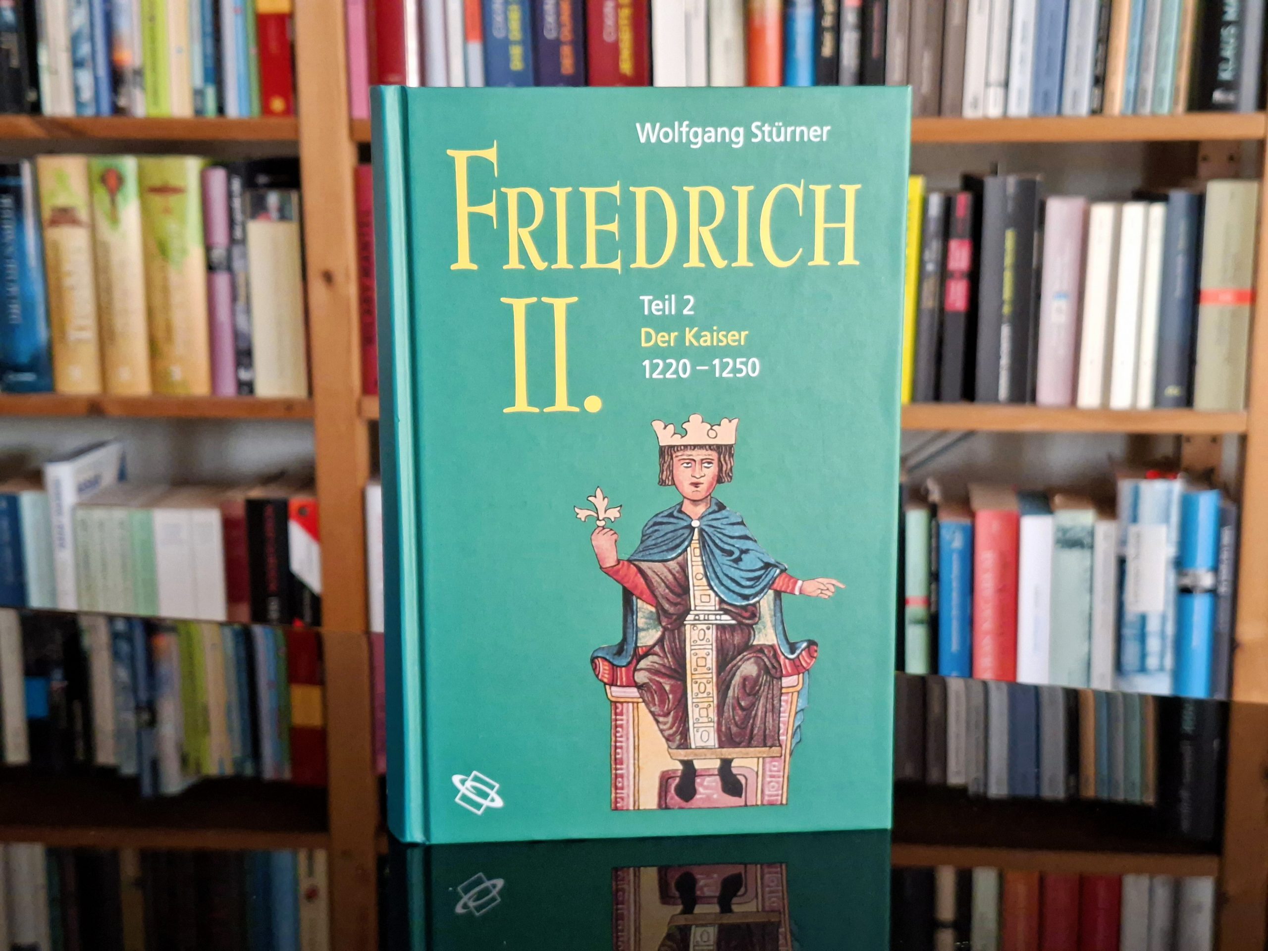 Ein Buch mit dem Titel 'Friedrich II., Teil 2: Der Kaiser 1220–1250' von Wolfgang Stürner, das vor einem Bücherregal steht. Das Cover zeigt eine Illustration von Kaiser Friedrich II. auf einem Thron mit Krone und Zepter.