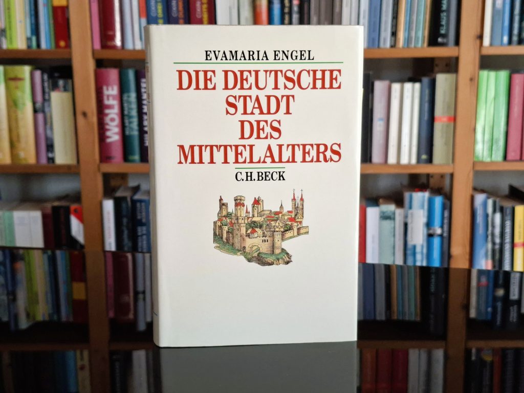 "Ein Buch mit dem Titel 'Die Deutsche Stadt des Mittelalters' von Evamaria Engel und C.H. Beck, das vor einem Regal mit zahlreichen weiteren Büchern steht. Das Buchcover zeigt eine Illustration einer mittelalterlichen Stadt mit Mauern, Türmen und Gebäuden.